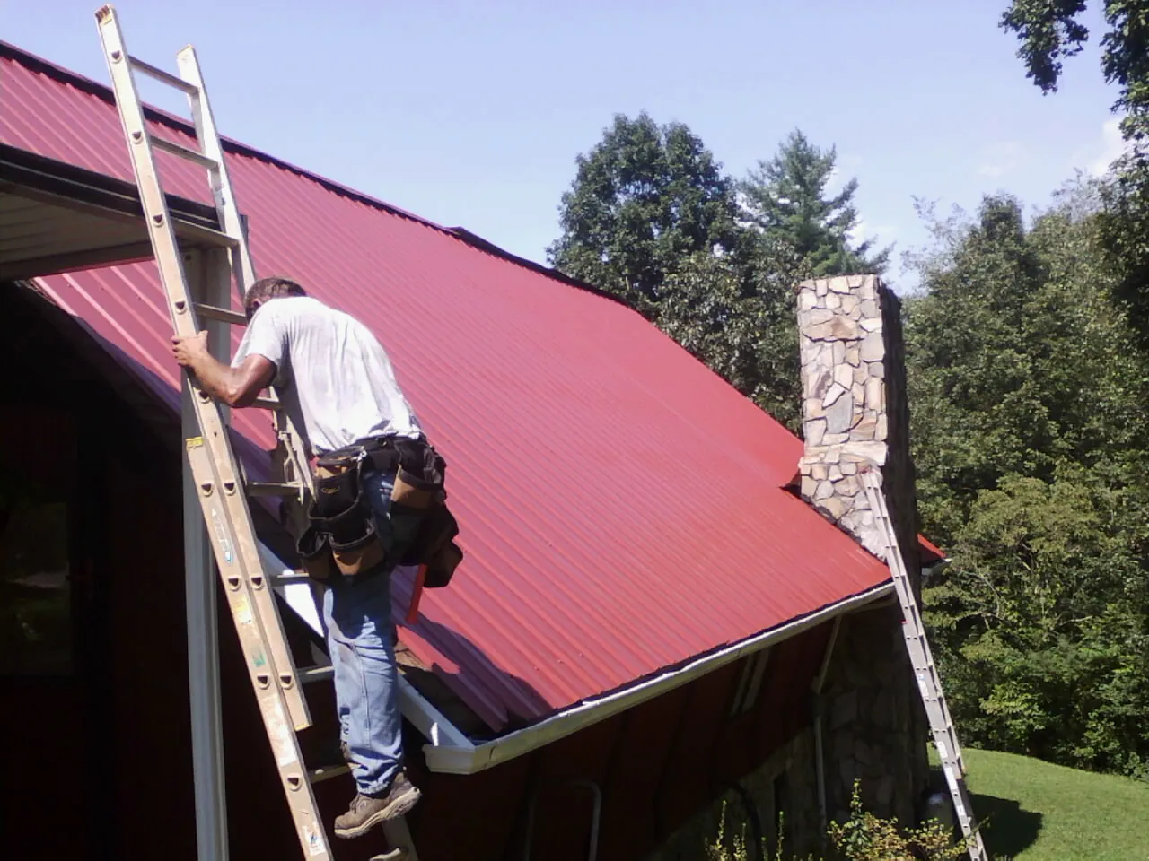 Roofer on a ladder beside a red metal roof and stone chimney — Soffit & Fascia Repair by Roofing Brothers in Manassas
